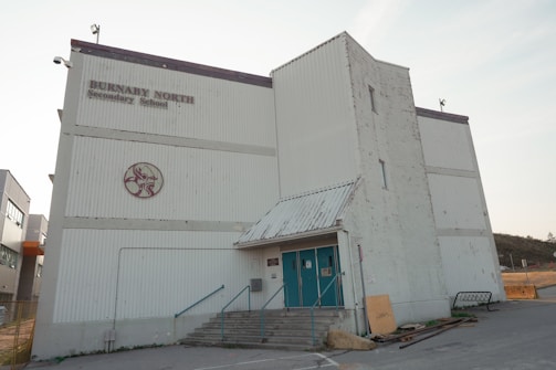 A large, white industrial-style building with a rectangular shape and corrugated metal exterior. There are faded letters reading 'Burnaby North Secondary School' and a circular emblem in red on the facade. The structure features a small overhang above the entrance, which has teal-colored double doors, accessible via a short set of concrete steps with metal railings. A few utility structures and equipment are scattered around, with signs of some wear and weathering on the walls.