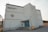 A large, white industrial-style building with a rectangular shape and corrugated metal exterior. There are faded letters reading 'Burnaby North Secondary School' and a circular emblem in red on the facade. The structure features a small overhang above the entrance, which has teal-colored double doors, accessible via a short set of concrete steps with metal railings. A few utility structures and equipment are scattered around, with signs of some wear and weathering on the walls.