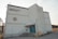 A large, white industrial-style building with a rectangular shape and corrugated metal exterior. There are faded letters reading 'Burnaby North Secondary School' and a circular emblem in red on the facade. The structure features a small overhang above the entrance, which has teal-colored double doors, accessible via a short set of concrete steps with metal railings. A few utility structures and equipment are scattered around, with signs of some wear and weathering on the walls.