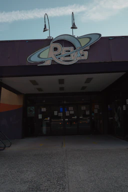 A building entrance with a large sign above the doors displaying the word 'REVS' designed with a circular orbit motif. Two lamps are mounted above the sign. The entrance features glass doors, and the surrounding areas are mostly concrete with dark-colored walls.