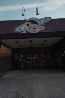 A building entrance with a large sign above the doors displaying the word 'REVS' designed with a circular orbit motif. Two lamps are mounted above the sign. The entrance features glass doors, and the surrounding areas are mostly concrete with dark-colored walls.