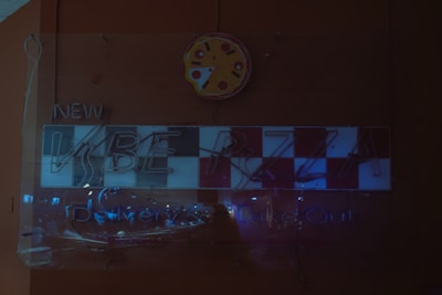 A dimly lit neon sign featuring a checkered pattern with the word 'PIZZA' partially visible and glowing faintly. Above the sign is a circular pizza slice graphic. The text 'NEW' and the words 'Delivery & Take Out' are also present but hard to read due to low lighting.