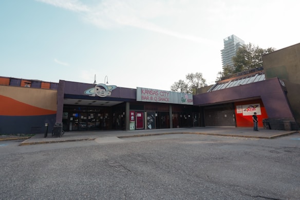 A building with signage for 'Kansas City Bar-B-Q Shack' and 'Revs Pizza' is shown. The structure has a modern yet slightly worn look with large glass doors and windows. There is a person standing towards the right side, and a tall, modern high-rise building is visible in the background. The sky is clear with a few clouds.