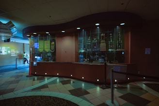 A dimly lit lobby area featuring a large curved trophy display case with several trophies inside. The floor is decorated with a geometric pattern of multicolored tiles, and banners hang on the wall near the trophies. In the background, a person is seated, partially obscured by shadows and a rope divider.