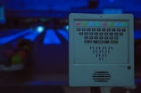 A dimly lit bowling alley with a control panel in the foreground. The panel has a QWERTY keyboard layout with colored buttons at the top and bowling pin icons at the bottom. Bowling lanes are visible in the blurred background, creating an ambient and slightly futuristic atmosphere.