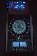 A digital dartboard is mounted within a dimly lit space, featuring a circular target labeled with numbered sections from 1 to 20. Below the target area, there are control panels with several buttons labeled P1 to P4, a start button, and additional controls. Above the dartboard, a screen displays a faint projection of an image, possibly of a game interface or other electronic display.