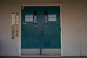 A set of teal double doors with room numbers 413 to 416 displayed. The doors are situated in a brick wall with light gray paint, showing signs of aging and some graffiti. On the left side, vertical signs read 'Technology' and 'Education'. The environment hints at an institutional or school setting.
