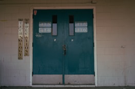 A set of teal double doors with room numbers 413 to 416 displayed. The doors are situated in a brick wall with light gray paint, showing signs of aging and some graffiti. On the left side, vertical signs read 'Technology' and 'Education'. The environment hints at an institutional or school setting.