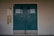 A set of teal double doors with room numbers 413 to 416 displayed. The doors are situated in a brick wall with light gray paint, showing signs of aging and some graffiti. On the left side, vertical signs read 'Technology' and 'Education'. The environment hints at an institutional or school setting.