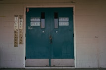A set of teal double doors with room numbers 413 to 416 displayed. The doors are situated in a brick wall with light gray paint, showing signs of aging and some graffiti. On the left side, vertical signs read 'Technology' and 'Education'. The environment hints at an institutional or school setting.