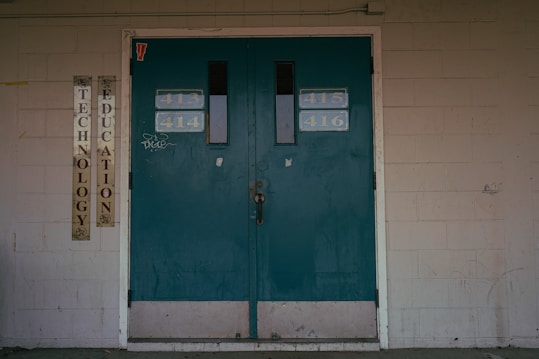 A set of teal double doors with room numbers 413 to 416 displayed. The doors are situated in a brick wall with light gray paint, showing signs of aging and some graffiti. On the left side, vertical signs read 'Technology' and 'Education'. The environment hints at an institutional or school setting.