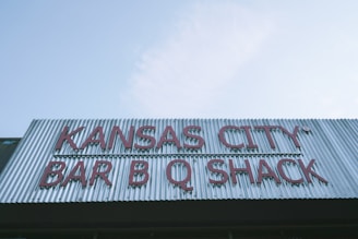 A vertical corrugated metal sign displays the words 'Kansas City Bar B Q Shack' in large red letters against a cloudy blue sky background.