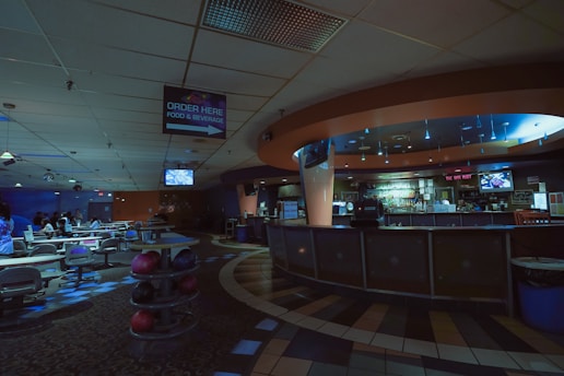 A dimly lit bowling alley features an empty bar area to the right and several lanes to the left. The ceiling is adorned with a mix of square tiles and light fixtures. A sign indicating 'Order Here' for food and beverages hangs from the ceiling. The space has a cozy, subdued ambiance with scattered seating and bowling balls visible.