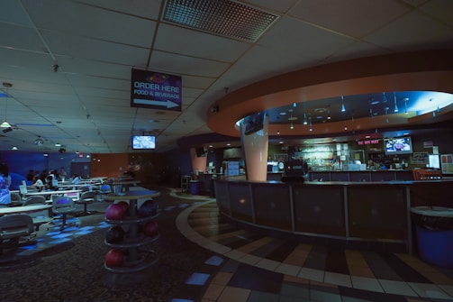 A dimly lit bowling alley features an empty bar area to the right and several lanes to the left. The ceiling is adorned with a mix of square tiles and light fixtures. A sign indicating 'Order Here' for food and beverages hangs from the ceiling. The space has a cozy, subdued ambiance with scattered seating and bowling balls visible.