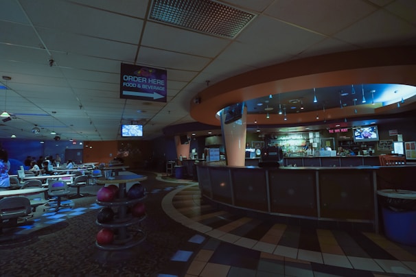 A dimly lit bowling alley features an empty bar area to the right and several lanes to the left. The ceiling is adorned with a mix of square tiles and light fixtures. A sign indicating 'Order Here' for food and beverages hangs from the ceiling. The space has a cozy, subdued ambiance with scattered seating and bowling balls visible.