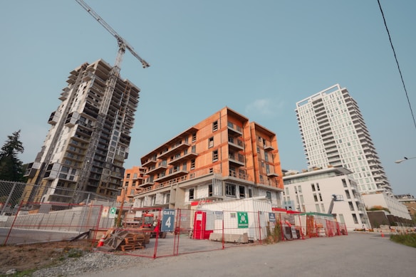 Multiple buildings at various stages of construction. There are cranes, scaffolding, and construction materials scattered around. The area is fenced off with 'Gate 4' prominently displayed on a sign. The buildings range from partially completed concrete structures to finished white high-rises. The sky is clear.