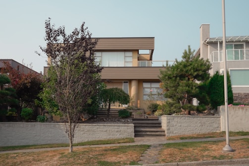 A modern two-story house with a minimalist style, featuring large windows and a flat roof. It is surrounded by well-maintained landscaping, including various shrubs and a tree in the front yard. A paved walkway leads up to the entrance.