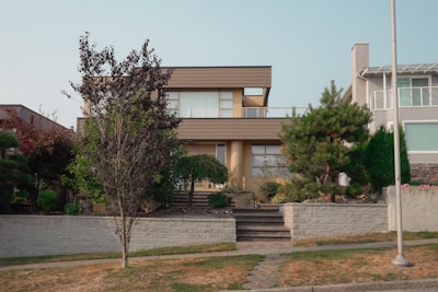 A modern two-story house with a minimalist style, featuring large windows and a flat roof. It is surrounded by well-maintained landscaping, including various shrubs and a tree in the front yard. A paved walkway leads up to the entrance.