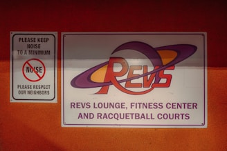 On a bright orange wall, two signs are mounted. The smaller sign to the left says 'PLEASE KEEP NOISE TO A MINIMUM, NOISE, PLEASE RESPECT OUR NEIGHBORS' with a graphic of a crossed-out noise icon. The larger sign on the right features the text 'REVS LOUNGE, FITNESS CENTER AND RACQUETBALL COURTS' with a stylized 'REVS' logo incorporating planetary rings.