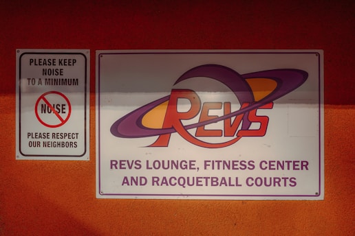 On a bright orange wall, two signs are mounted. The smaller sign to the left says 'PLEASE KEEP NOISE TO A MINIMUM, NOISE, PLEASE RESPECT OUR NEIGHBORS' with a graphic of a crossed-out noise icon. The larger sign on the right features the text 'REVS LOUNGE, FITNESS CENTER AND RACQUETBALL COURTS' with a stylized 'REVS' logo incorporating planetary rings.