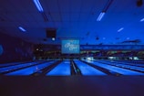 A dimly lit bowling alley with multiple lanes illuminated by blue lights. The ceiling features long light fixtures casting a faint glow. A large screen at the end of the lanes displays text. Walls are decorated with abstract patterns creating a futuristic or cosmic ambiance.
