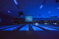A dimly lit bowling alley with multiple lanes illuminated by blue lights. The ceiling features long light fixtures casting a faint glow. A large screen at the end of the lanes displays text. Walls are decorated with abstract patterns creating a futuristic or cosmic ambiance.