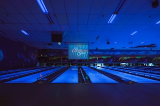 A dimly lit bowling alley with multiple lanes illuminated by blue lights. The ceiling features long light fixtures casting a faint glow. A large screen at the end of the lanes displays text. Walls are decorated with abstract patterns creating a futuristic or cosmic ambiance.