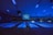 A dimly lit bowling alley with multiple lanes illuminated by blue lights. The ceiling features long light fixtures casting a faint glow. A large screen at the end of the lanes displays text. Walls are decorated with abstract patterns creating a futuristic or cosmic ambiance.