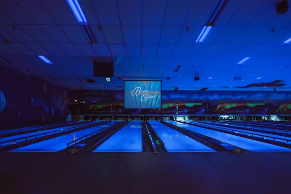 A dimly lit bowling alley with multiple lanes illuminated by blue lights. The ceiling features long light fixtures casting a faint glow. A large screen at the end of the lanes displays text. Walls are decorated with abstract patterns creating a futuristic or cosmic ambiance.
