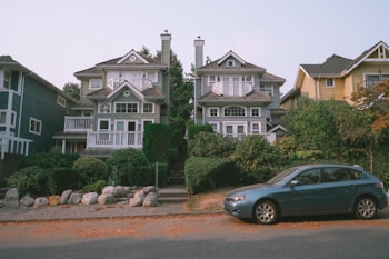 Two large, identical, three-story houses with white trim and extensive glass windows are at the center of the image, separated by a staircase lined with greenery. A blue car is parked on the road in the foreground. There are well-maintained bushes and trees surrounding the homes, adding a lush green appearance. The sky overhead is clear with a hint of light blue. Nearby houses are visible, with one distinct yellow house to the right.