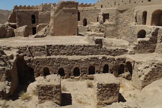 A historical site featuring ancient stone and mud-brick structures. The architecture includes arched doorways and windows, with multi-level stone walls set against a mountainous backdrop. The sun casts harsh shadows, suggesting a desert environment.