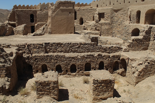 A historical site featuring ancient stone and mud-brick structures. The architecture includes arched doorways and windows, with multi-level stone walls set against a mountainous backdrop. The sun casts harsh shadows, suggesting a desert environment.