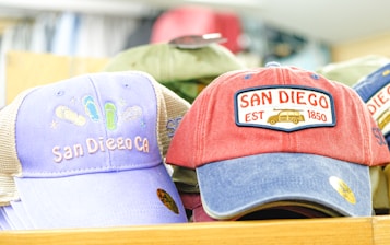 A collection of colorful caps is displayed, each featuring embroidered designs related to San Diego. The caps showcase casual designs with one reading 'San Diego CA' and another featuring a red, white, and blue color scheme with 'San Diego EST 1850' and an illustration of a van.