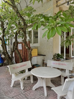 A small outdoor seating area features a white concrete table with two benches. The benches have red text, one reading 'THE CHILL GOURMET' and the other 'THUẬN HẢI HOTEL'. Lush green tree branches with broad leaves hang overhead, casting shadows on the tiled ground.