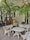 A small outdoor seating area features a white concrete table with two benches. The benches have red text, one reading 'THE CHILL GOURMET' and the other 'THUẬN HẢI HOTEL'. Lush green tree branches with broad leaves hang overhead, casting shadows on the tiled ground.