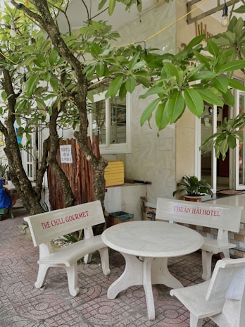 A small outdoor seating area features a white concrete table with two benches. The benches have red text, one reading 'THE CHILL GOURMET' and the other 'THUẬN HẢI HOTEL'. Lush green tree branches with broad leaves hang overhead, casting shadows on the tiled ground.