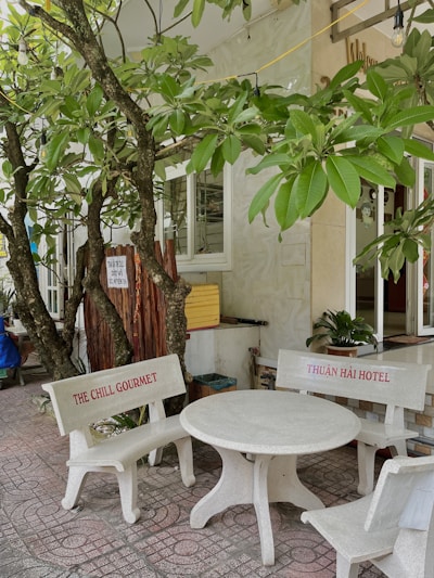 A small outdoor seating area features a white concrete table with two benches. The benches have red text, one reading 'THE CHILL GOURMET' and the other 'THUẬN HẢI HOTEL'. Lush green tree branches with broad leaves hang overhead, casting shadows on the tiled ground.