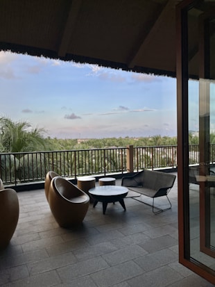 An outdoor terrace with modern furniture including round chairs, small tables, and a bench. The area is tiled and overlooks a lush green landscape with palm trees under a partially cloudy sky. The terrace is partially covered by a dark roof and bordered by a railing.
