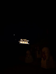 A brightly lit traditional building is visible against the dark night sky, accompanied by the faint glow of a crescent moon. In the foreground, two stone lion statues are partially illuminated, adding a sense of majesty and antiquity to the scene.