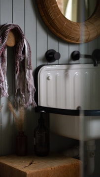 A cozy bathroom corner with a rustic aesthetic featuring a round wicker-framed mirror above a vintage-style sink with black fixtures. A striped towel is draped over a wooden peg on the wall, accompanied by a small vase with dried plants and a dark glass bottle on a wooden surface.