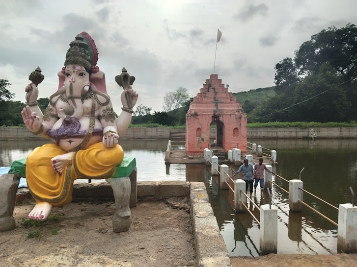 A large statue of the Hindu deity Ganesha sits on a raised platform beside a body of water. Ganesha, with an elephant head, is adorned with a traditional headdress and jewelry and is seated in a lotus position, holding objects in multiple arms. In the background, a pink temple structure stands partially submerged in water. Two people are walking on a wet pathway that leads to the temple, surrounded by lush greenery and cloudy skies.