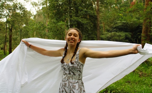 A young person with braided hair is joyfully holding a large white sheet in a lush green forest. The trees and foliage in the background create a serene natural setting.