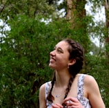 A young person with braided hair is smiling and looking upward. They are surrounded by lush greenery, with tall trees in the background, suggesting a natural, outdoor setting.
