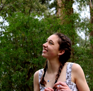 A young person with braided hair is smiling and looking upward. They are surrounded by lush greenery, with tall trees in the background, suggesting a natural, outdoor setting.