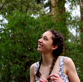 A young person with braided hair is smiling and looking upward. They are surrounded by lush greenery, with tall trees in the background, suggesting a natural, outdoor setting.