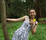 A young person with braided hair is smiling while holding a yellow flower. They are leaning against a tree in a lush forest setting with green grass and small white flowers scattered around.