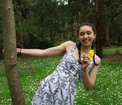 A young person with braided hair is smiling while holding a yellow flower. They are leaning against a tree in a lush forest setting with green grass and small white flowers scattered around.