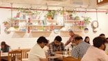 A cozy indoor café scene with several people sitting at wooden tables, engaging in different activities like checking their phones or chatting. The white brick wall is adorned with shelves filled with books, potted plants, and decorative items. Warm pendant lights hang from the ceiling, contributing to the inviting ambiance.