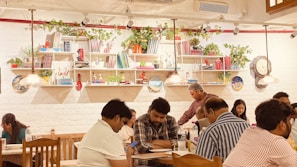 A cozy indoor café scene with several people sitting at wooden tables, engaging in different activities like checking their phones or chatting. The white brick wall is adorned with shelves filled with books, potted plants, and decorative items. Warm pendant lights hang from the ceiling, contributing to the inviting ambiance.