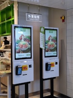 Two digital kiosks for placing takeout orders, displaying images of food items. They are located in a restaurant setting with visible payment interfaces and a sign above indicating 'Takeout Order' in both English and Chinese.